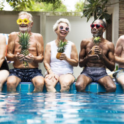 Group of diverse senior adults sitting at poolside holding pineapples together proving aging is not boring. Q4fit.com
